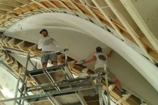 Two men painting a curved ceiling in a spacious room with natural light coming in from a large window.